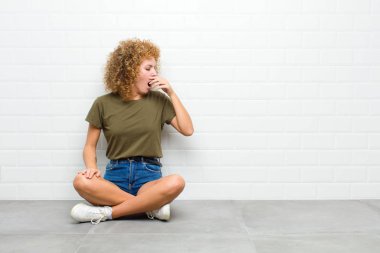 Genç afro kadın sabahın erken saatlerinde uyanıyor, uykulu görünüyor, yorgun ve sıkkın bir şekilde yerde oturuyor.