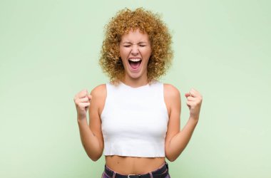 Genç afro kadın son derece mutlu ve şaşırmış görünüyor, başarıyı kutluyor, bağırıyor ve yeşil duvara zıplıyor.