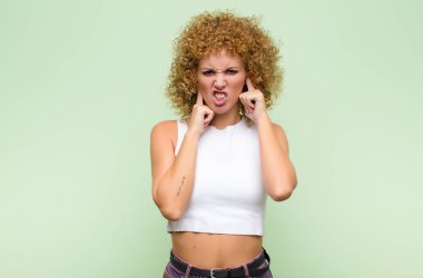 Genç afro kadın sinirli, stresli ve sinirli görünüyor, her iki kulağı da sağır edici bir gürültü, ses veya yüksek sesle kaplayan yeşil duvara karşı müzik