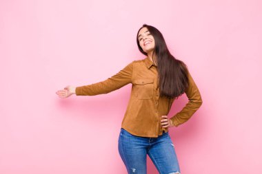 Genç, güzel bir kadın kendini mutlu ve neşeli hissediyor, gülümsüyor ve seni hoş karşılıyor, pembe duvara karşı dostça bir jestle içeri davet ediyor.