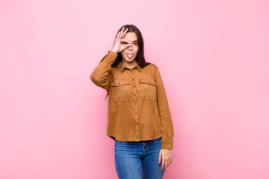 Genç ve güzel bir kadın mutlu bir şekilde gülümsüyor, şaka yapıyor ve gözetleme deliğinden bakıyor, pembe duvara karşı sırları gözetliyor.