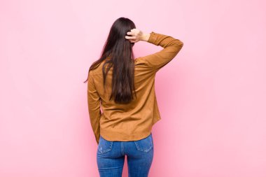 Genç güzel bir kadın düşünüyor ya da şüphe ediyor, kafasını kaşıyor, şaşkın ve şaşkın hissediyor, pembe duvarın arkasında ya da arkasında.