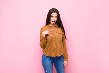 Genç ve güzel bir kadın şok olmuş ve şaşırmış görünüyor, ağzı sonuna kadar açık, pembe duvara karşı kendini işaret ederek.