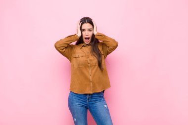 Genç ve güzel bir kadın hoş olmayan bir şekilde şok olmuş, korkmuş ya da endişeli görünüyor, ağzı sonuna kadar açık ve pembe duvara karşı iki kulağını da kapatıyor.