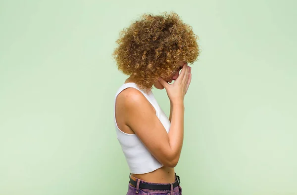 若いアフロの女性は手で目を覆い絶望の悲しい欲求不満そうな表情で泣き緑の壁に対するサイドビュー — ストック写真