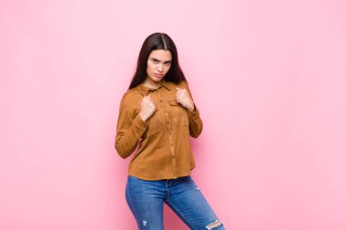 Kendine güvenen, sinirli, güçlü ve agresif görünen genç bir kadın. Yumrukları boks pozisyonunda pembe duvara karşı savaşmaya hazır.