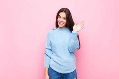 Genç, güzel bir kadın mutlu ve neşeyle gülümsüyor, el sallıyor, seni selamlıyor, ya da pembe duvara veda ediyor.