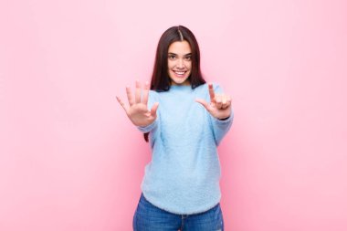 Genç, güzel bir kadın gülümsüyor ve arkadaş canlısı görünüyor. Elleri önde, pembe duvara doğru geri sayarak yedi ya da yedinci numarayı gösteriyor.