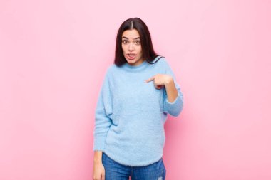 Genç, güzel bir kadın mutlu, gururlu ve şaşırmış görünüyor. Neşeli bir şekilde kendini gösteriyor. Kendinden emin ve pembe duvara karşı kibirli hissediyor.