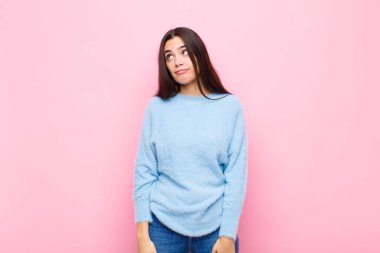 Genç ve güzel bir kadın şaşkın ve şaşkın görünüyor, merak ediyor veya bir sorunu çözmeye çalışıyor veya pembe duvara karşı düşünüyor.