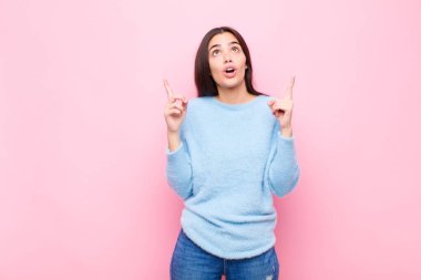 Genç ve güzel bir kadın huşu içinde ve ağzı açık bir şekilde pembe duvara doğru şaşkın ve şaşkın bir bakışla yukarıya bakıyor.