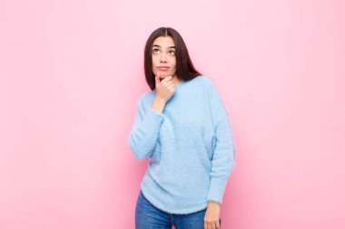 Genç, güzel bir kadın düşünüyor, kuşkulu ve şaşkın hissediyor, farklı seçeneklerle, pembe duvara karşı hangi kararı vereceğini merak ediyor.