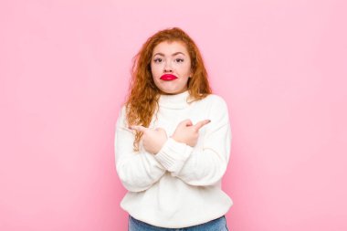 Genç kızıl saçlı kadın şaşkın ve şaşkın görünüyor, güvensiz ve pembe duvara karşı şüpheyle zıt yönleri işaret ediyor.