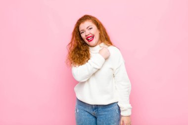 Genç kızıl saçlı kadın mutlu, pozitif ve başarılı hissediyor, meydan okumayla karşılaştığında motive oluyor veya pembe duvara karşı iyi sonuçları kutluyor.