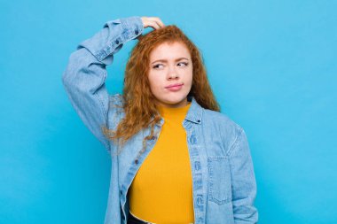 Genç kızıl saçlı kadın şaşkın ve kafası karışmış hissediyor. Kafasını kaşıyor ve mavi duvara bakıyor.