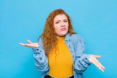 Genç kızıl saçlı kadın şaşkın, kafası karışmış ve stresli görünüyor, farklı seçenekler arasında merak ediyor, mavi duvara karşı kendini güvensiz hissediyor.