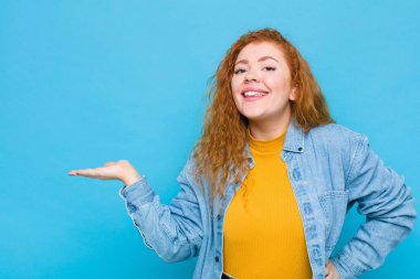 Genç kızıl saçlı kadın gülümsüyor, kendinden emin, başarılı ve mutlu hissediyor. Mavi duvara karşı fotokopi alanında fikir veya konsept gösteriyor.