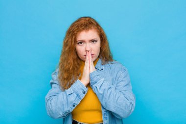 Genç kızıl saçlı kadın endişeli, umutlu ve dindar hissediyor. Avuçlarına bastırılmış, mavi duvara karşı af diliyor.