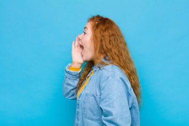 Genç kızıl saçlı kadın profili, mutlu ve heyecanlı görünüyor, bağırıyor ve mavi duvara karşı boşluğu kopyalamak için çağırıyor.