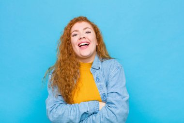 Genç kızıl saçlı kadın mutlu, gururlu ve memnun görünüyor. Kollarını mavi duvara dolamış gülümsüyor.