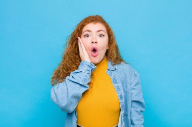Genç kızıl saçlı kadın şok olmuş ve şaşkınlık içinde. Mavi duvara karşı ağzı sonuna kadar açık bir şekilde inanmayarak el ele tutuşuyor.