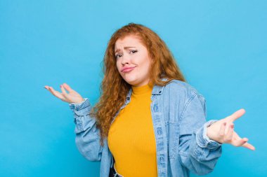 Genç kızıl saçlı kadın aptal, deli, kafası karışık, şaşkın bir ifadeyle omuz silkiyor, sinirlenmiş ve mavi duvara karşı bilgisiz hissediyor.