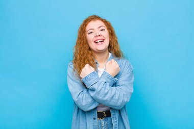 Genç kızıl saçlı, neşeli bir şekilde gülümseyen ve kollarını kavuşturup kollarını kavuşturan, mavi duvara karşı mutlu ve pozitif hisseden bir kadın.