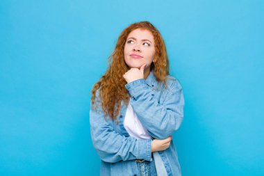 Genç kızıl saçlı kadın düşünceli, meraklı ya da hayal kuruyor, hayal kuruyor ve mavi duvara karşı mekanı kopyalamak için yukarı bakıyor.