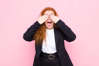 Genç kızıl saçlı kadın gülümsüyor ve mutlu hissediyor, iki eliyle gözlerini kapatıyor ve pembe duvara karşı inanılmaz bir sürpriz bekliyor.