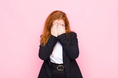 Genç kızıl saçlı kadın üzgün, hayal kırıklığına uğramış, gergin ve depresif, yüzünü iki eliyle kapatıyor, pembe duvara karşı ağlıyor.