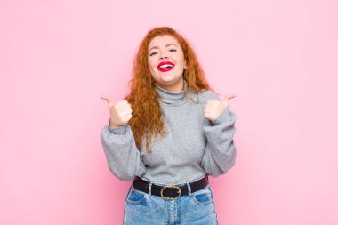 Genç kızıl saçlı kadın neşeyle gülümsüyor ve mutlu görünüyor, kaygısız ve pozitif hissediyor iki başparmağı pembe duvara dayalı