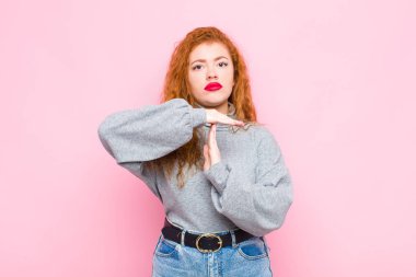 Genç kızıl saçlı kadın ciddi, sert, kızgın ve hoşnutsuz görünüyor. Pembe duvara karşı zaman geçirme işareti yapıyor.