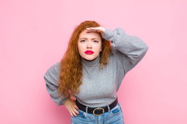 Genç kızıl saçlı kadın şaşkın şaşkın ve şaşkın bakıyor, elini alnına uzatmış uzaklara bakıyor, pembe duvara bakıyor ya da onu arıyor.