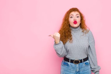 Genç kızıl saçlı kadın inançsızlıktan şaşkın şaşkın bakıyor, yan taraftaki objeyi işaret ediyor ve diyor ki vay be, pembe duvara karşı inanılmaz.