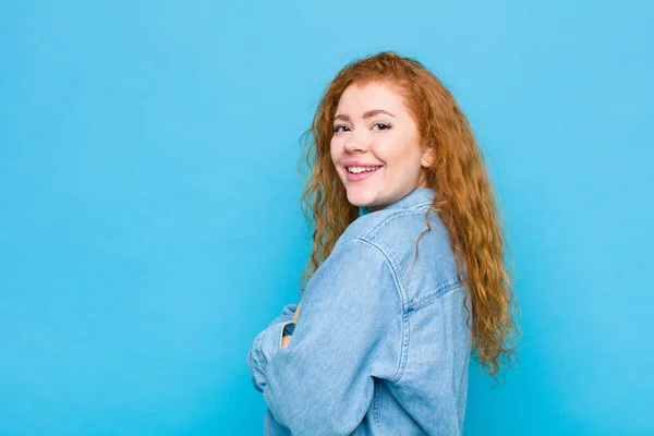 young red head woman smiling to camera with crossed arms and a happy, confident, satisfied expression, lateral view against blue wall