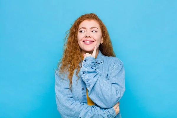 young red head woman smiling happily and daydreaming or doubting, looking to the side against blue wall