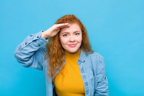 young red head woman looking bewildered and astonished, with hand over forehead looking far away, watching or searching against blue wall