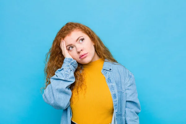 young red head woman feeling bored, frustrated and sleepy after a tiresome, dull and tedious task, holding face with hand against blue wall