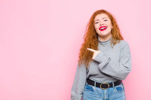 young red head woman smiling cheerfully, feeling happy and pointing to the side and upwards, showing object in copy space against pink wall