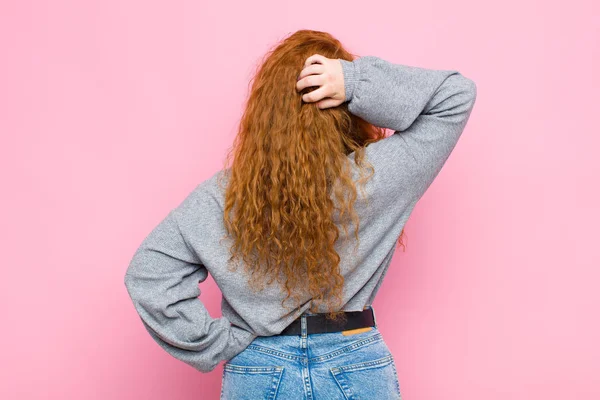 young red head woman thinking or doubting, scratching head, feeling puzzled and confused, back or rear view against pink wall