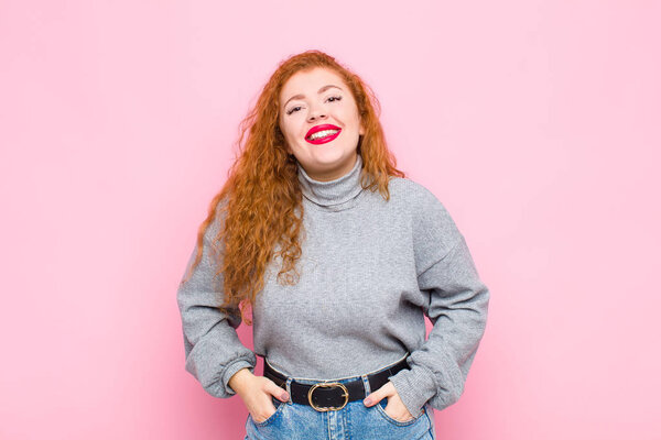 young red head woman smiling cheerfully and casually with a positive, happy, confident and relaxed expression against pink wall