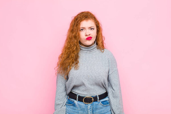 young red head woman feeling confused and doubtful, wondering or trying to choose or make a decision against pink wall