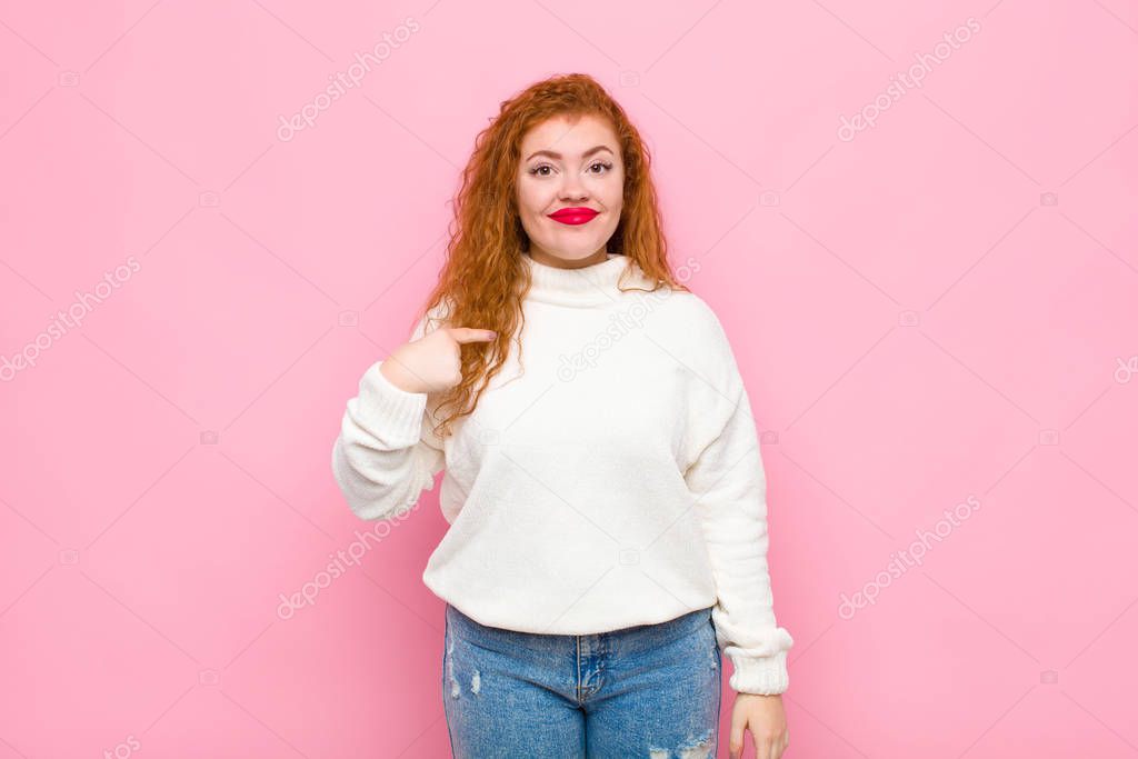 young red head woman looking proud, confident and happy, smiling and pointing to self or making number one sign against pink wall