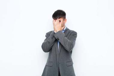 Genç yakışıklı adam elleriyle yüzünü kapatıyor, parmaklarının arasından şaşkın bir ifadeyle bakıyor ve beyaz duvara bakıyor.