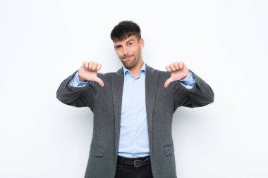 Genç yakışıklı adam üzgün, hayal kırıklığına uğramış ya da kızgın görünüyor, anlaşmazlık içinde başparmaklarını gösteriyor, beyaz duvara karşı hayal kırıklığına uğramış hissediyor.