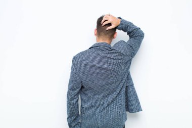 Genç, yakışıklı bir adam bilgisiz ve kafası karışık, bir çözüm düşünüyor, eli kalçasında, diğeri kafasında, beyaz duvara karşı dikiz manzaralı.