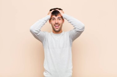 young handsome man raising hands to head, open-mouthed, feeling extremely lucky, surprised, excited and happy against warm wall