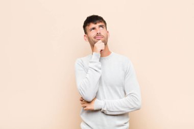 young handsome man thinking, feeling doubtful and confused, with different options, wondering which decision to make against warm wall
