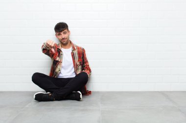young handsome man feeling cross, angry, annoyed, disappointed or displeased, showing thumbs down with a serious look sitting on cement floor