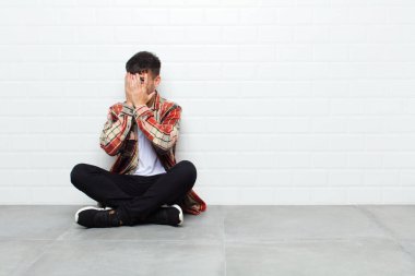 young handsome man covering face with hands, peeking between fingers with surprised expression and looking to the side sitting on cement floor